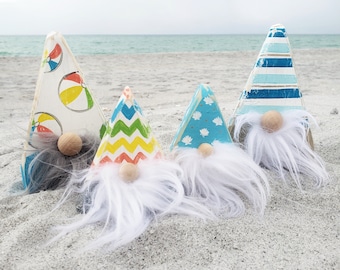May include: Four wooden gnome figurines with white, blue, and yellow paint. Each gnome has a different design and a white beard. The gnomes are sitting on a sandy beach.