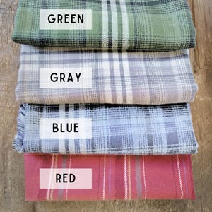 May include: Four folded plaid fabric samples in green, gray, blue, and red. Each fabric has a white rectangular label with the color name printed in a bold font. The fabrics are stacked on a wooden surface.