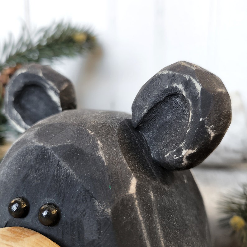 May include: Close-up of a black wooden bear sculpture with visible wood grain and white accents. The bear has two black bead eyes and two large ears. The sculpture is a rustic, handcrafted item.