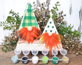 May include: Two unfinished wooden gnome figures with red and orange fur beards. The gnomes are decorated with green and gold plaid and shamrock patterns. The figures are sitting on a white wooden shelf with six small paint pots in front of them. The paint pots are green, black, white, gold, and two shades of green.