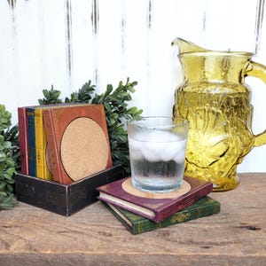 May include: Set of six book-shaped coasters with cork tops. The coasters are stacked in a black wooden holder. A glass of ice water and a yellow glass pitcher are on a wooden table.