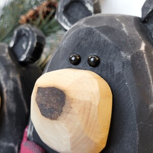 May include: Close-up of a wooden bear head with black painted fur and a natural wood-colored snout. The bear has black bead eyes and a carved appearance. The image suggests a rustic, handcrafted decorative item.