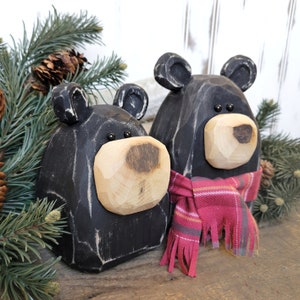 May include: Two black wooden bear figurines with light-colored wooden snouts and small black eyes. One bear wears a red plaid scarf. The bears are surrounded by pine needles and pine cones, creating a rustic, woodland theme.