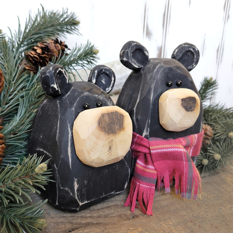 May include: Two black wooden bear figurines with light-colored wooden snouts and small black eyes. One bear wears a red plaid scarf. The bears are surrounded by pine needles and pine cones, creating a rustic, woodland theme.