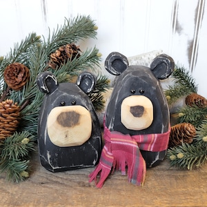 May include: Two black wooden bear figurines with light brown snouts and ears. One bear wears a red and pink plaid scarf. The bears are surrounded by pine branches and pine cones, creating a rustic, woodland theme.