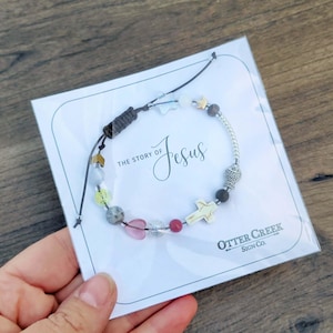 Pu&ograve; includere: Un braccialetto di perline con un ciondolo a forma di croce e la scritta "The Story of Jesus" su una carta bianca. Il braccialetto presenta varie perline colorate e un ciondolo a forma di pesce. La carta ha anche il logo "Otter Creek Sign Co.".