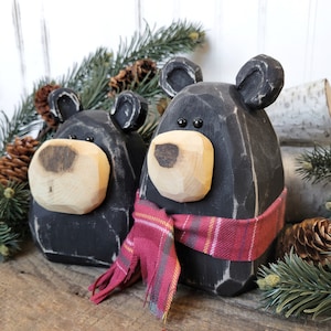 May include: Two black wooden bear figurines with light-colored wooden snouts and ears. One bear wears a red plaid scarf. The bears are on a wooden surface with pine needles and pine cones in the background.