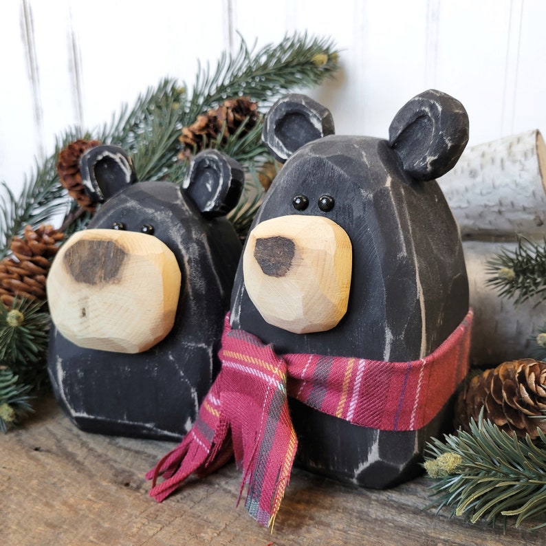 May include: Two black wooden bear figurines with light-colored wooden snouts and ears. One bear wears a red plaid scarf. The bears are on a wooden surface with pine needles and pine cones in the background.