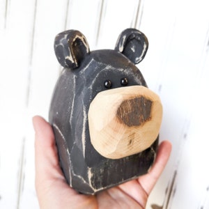May include: A hand-carved wooden bear head with a black painted body and a natural wood-colored snout. The bear has small, black bead eyes and a rustic, distressed finish, perfect for home decor.