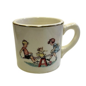 May include: Cream-colored ceramic mug with a gold rim and a curved handle. The mug features a colorful illustration of three children playing on a seesaw. A red seesaw and a red ball are also visible in the image.