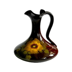 Puede incluir: Jarra de cerámica marrón oscuro pintada a mano con un diseño floral amarillo. La jarra tiene un asa curva y un pico acampanado. El diseño floral presenta una gran flor amarilla con un centro rojo y hojas verdes.