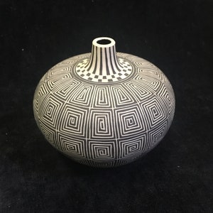 May include: A round, black and white ceramic vase with a geometric pattern. The vase has a narrow neck with vertical black stripes. The body of the vase is covered in a repeating square design. The vase is a decorative item.