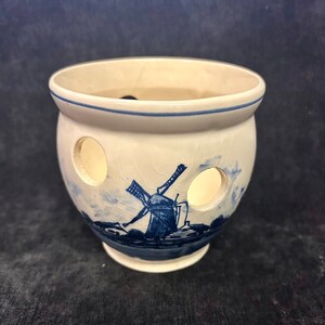 May include: A ceramic onion pot with a blue and white windmill design. The pot has a round shape with three circular holes and a blue rim. The windmill scene is painted on the front, with a blue base and a white background.