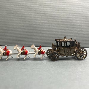 May include: A miniature antique carriage pulled by five white horses, each with a rider in a red coat. The carriage is bronze-colored with intricate detailing. The horses are connected to the carriage by a silver bar.