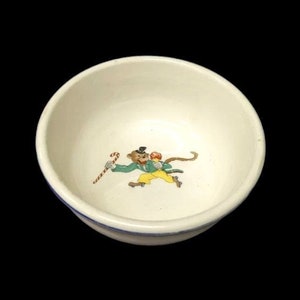 May include: A small, off-white ceramic bowl with a blue rim. The interior features a colorful illustration of a monkey holding a candy cane. The monkey is wearing a green jacket, yellow pants, and a black hat.