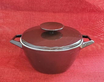 Op de afbeelding: Een donkerbruine braadpan met deksel, zwarte handvatten en een witte rand. Het deksel heeft een kleine, ronde handgreep. De pan staat tegen een rode achtergrond.