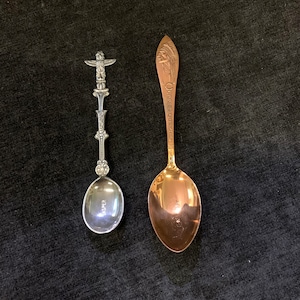 Könnte beinhalten: Zwei dekorative Löffel. Einer ist silberfarben mit einem Totempfahl-Design am Griff und dem Wort "CASPER" auf der Schale. Der andere ist goldfarben mit einem eingravierten indianischen Profil und Text auf dem Griff.