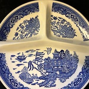 Pode incluir: Um prato branco dividido com um design azul padrão Willow. O prato é dividido em três seções. O design azul apresenta uma paisagem com edifícios, árvores e um barco. A borda tem uma borda decorativa.