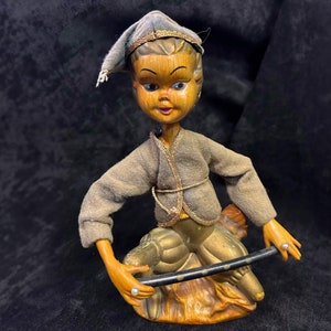 May include: A vintage wooden doll with a painted face, wearing a grey outfit and hat. The doll is posed with a black rod, and has gold-coloured accents. The doll's hands are painted orange, and it is sitting on a gold-coloured base.