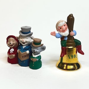 May include: A collection of Christmas figurines. Three animal figures in red, blue, and green outfits are holding books. A gold bell with a gnome figure is also present. The year "1991" is printed on the bell and book.