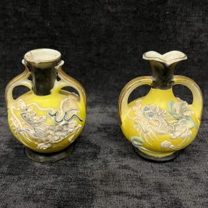 May include: Two small, yellow vases with black and gold accents. Each vase features a detailed, silver-colored dragon design. The vases have a unique shape with handles and a flared top, perfect for decorative purposes.