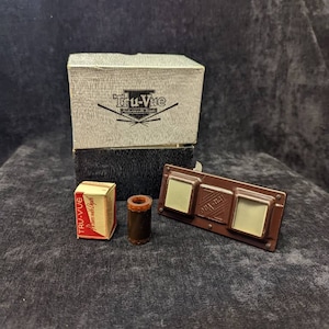Puede incluir: Una colección de visores y accesorios Tru-Vue vintage. Los artículos incluyen un visor, una caja pequeña, un objeto cilíndrico y una caja de imágenes Tru-Vue con profundidad. El visor es marrón con dos ventanas de visualización cuadradas.