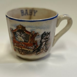 May include: Antique ceramic baby cup with a blue rim and handle. The cup features a charming illustration of kittens in a carriage and dancing. The word "BABY" is printed in blue on the cup's rim. A vintage collectible.