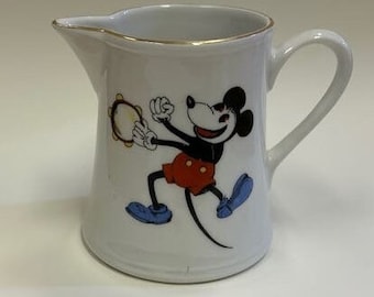 May include: White ceramic creamer with a gold rim and a handle. The creamer features a colorful illustration of Mickey Mouse playing a tambourine. Mickey is wearing red shorts, blue shoes, and a black shirt.