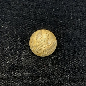 Puede incluir: Botón redondo, color dorado, con un diseño en relieve que representa un águila con las alas extendidas y un escudo. El botón está sobre un fondo oscuro y texturizado, resaltando los detalles del botón.