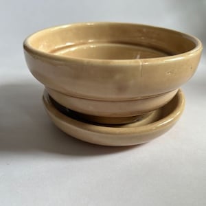 May include: A beige ceramic bowl with a matching saucer. The bowl has a rounded shape with a slightly flared rim. The saucer is a smaller, shallow dish. Both pieces have a glossy finish.