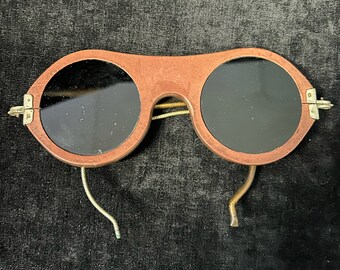 May include: Vintage sunglasses featuring dark circular lenses within a reddish-brown wooden frame. The frame has a distinctive shape, with a bridge and hinges. The temples are crafted from thin, gold-coloured metal wire. A classic accessory.