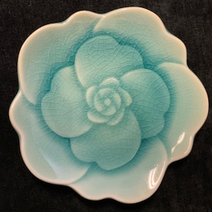 Op de afbeelding: Een keramische schaal in de vorm van een bloem, in tinten turquoise en wit. De schaal heeft een craquelé-glazuur en een geschulpte rand. Het midden van de bloem is verhoogd en de bloemblaadjes zijn gedetailleerd.