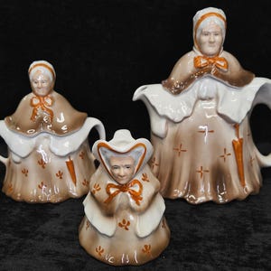 May include: A set of three ceramic figurines, including a teapot, a creamer, and a sugar bowl, all in the shape of a woman. The figurines are primarily brown and white, with orange accents. The teapot has a handle and spout.
