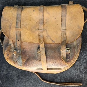 May include: A vintage leather satchel in shades of brown, featuring a curved flap and multiple leather straps with buckles. The bag shows signs of wear, with a long strap extending from the bottom.