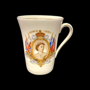Op de afbeelding: Crèmekleurige herdenkingsmok met een portret van koningin Elizabeth II, met de tekst "June 2 1953" en "Coronation Elizabeth". De mok heeft een gouden rand en oor en bevat vlaggen.