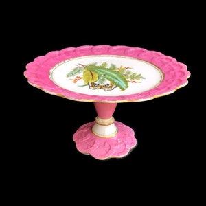 Może przedstawiać: Różowy stojak na ciasto z falistą krawędzią i kwiatowym wzorem na talerzu. Talerz ma biały środek z zielono-żółtą ilustracją botaniczną. Stojak ma różową podstawę i trzon z złotym wykończeniem.