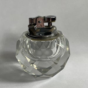 Puede incluir: Un encendedor de cristal transparente y facetado con una parte superior de metal plateado. El encendedor se asienta sobre una base redonda de cristal transparente con un patrón de corte de diamante. El encendedor es de estilo vintage.