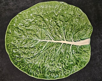 Puede incluir: Una fuente de cerámica verde con forma de hoja de col. La fuente tiene una superficie texturizada con vetas detalladas y un tallo de color crema. El diseño es una representación realista de una hoja de col.