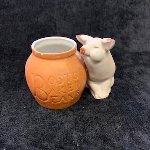May include: A ceramic Boston Baked Beans jar with a smiling pig figurine. The jar is a light orange color with the words "Boston Baked Beans" embossed on the side. The pig is white with pink accents on its ears and snout.