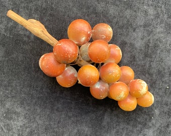 May include: A cluster of faux grapes with a wooden stem. The grapes are a mix of orange and brown, with some showing white spots. The stem is a light brown color. The grapes are arranged in a bunch.