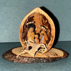 Può includere: Un presepe in miniatura scolpito all'interno di un guscio di noce. Le figure sono dipinte con dettagli, tra cui aloni e abiti. La noce è montata su una piccola base di legno rotonda, con un pezzo di legno davanti.