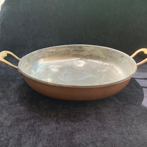 Puede incluir: Una sartén de color cobre con interior plateado y dos asas de latón. La sartén es de forma ovalada y parece ser vintage. Las asas son curvas y están unidas a los lados de la sartén. La sartén está sobre una superficie oscura.