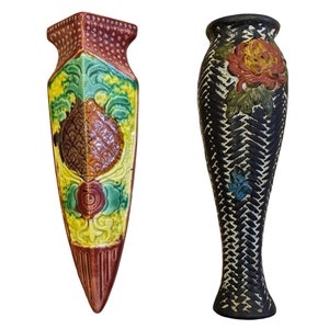 May include: Two decorative ceramic vases. One is a tall, black vase with a textured pattern and a painted flower. The other is a colorful, shield-shaped vase with a pineapple design and floral accents. Both vases are on a white background.
