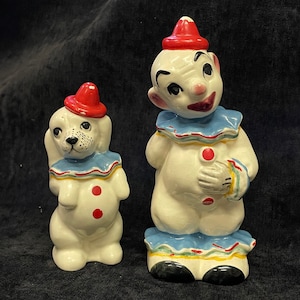 Könnte beinhalten: Zwei Clownfiguren aus Keramik. Eine ist ein Hund mit rotem Hut und blauem Kragen. Die andere ist ein Clown mit rotem Hut, blauem Kragen und schwarzen Schuhen. Beide haben rote Knöpfe und sind im Vintage-Stil bemalt.