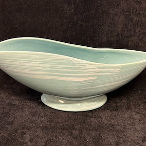 Può includere: Una ciotola da portata in ceramica azzurra con base a piedistallo. La ciotola ha una forma asimmetrica unica con un bordo curvo e striature orizzontali bianche. Un pezzo decorativo.