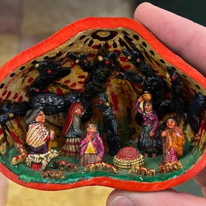 Può includere: Diorama in miniatura dipinto a mano, incorniciato in una forma a conchiglia rossa. La scena raffigura figure in abiti tradizionali, alberi e un cesto, il tutto in colori vivaci. L'interno presenta un motivo. Tenuto in mano.