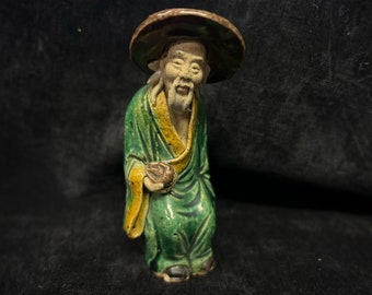 May include: A ceramic figurine of an elderly man wearing a wide-brimmed hat and a long green robe with yellow trim. The figure is holding a small bowl and has a serene expression. The figurine has a weathered appearance, suggesting age and craftsmanship.