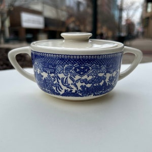 Puede incluir: Un azucarero de cerámica azul y blanco con tapa. El cuenco presenta un diseño Willow y dos asas. La tapa es de color crema. El azucarero está sobre una superficie blanca.