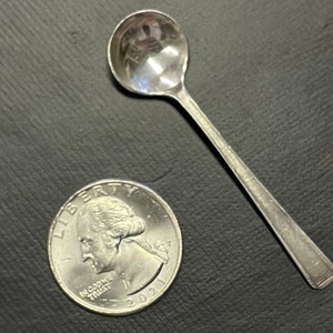 Puede incluir: Una pequeña cuchara de plata con un cuenco redondeado y un mango largo y delgado. Junto a la cuchara hay una moneda de veinticinco centavos estadounidense. La cuchara probablemente se utiliza para servir o medir pequeñas cantidades de alimentos o ingredientes.