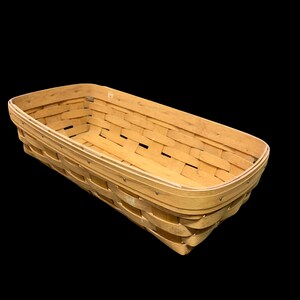 Puede incluir: Una cesta rectangular de mimbre marrón claro con un borde redondeado. La cesta está hecha de tiras de madera entrelazadas, con pequeños remaches metálicos que aseguran el borde. La cesta está vacía y se encuentra sobre un fondo negro.